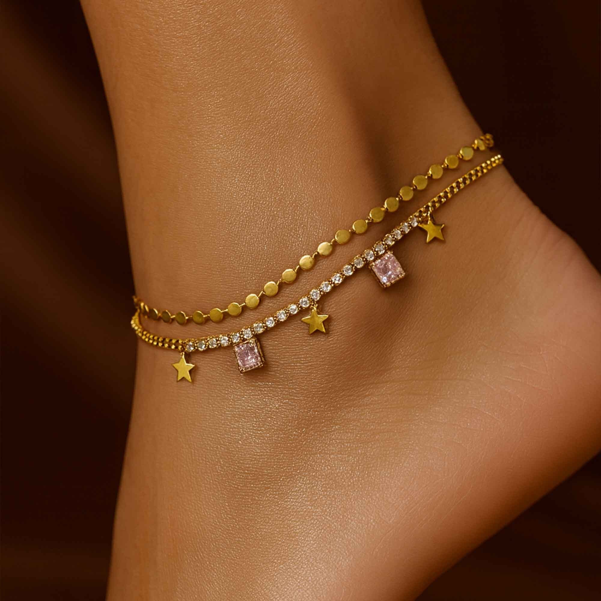 Gold anklets with star and gemstone charms on a person's ankle against a brown background