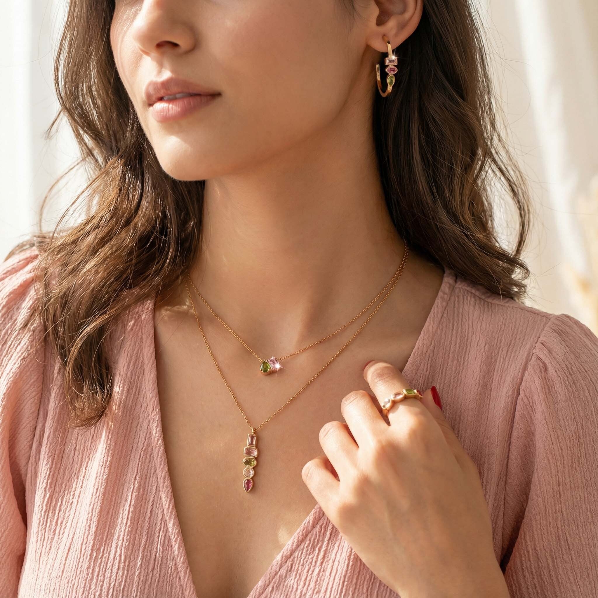 Woman wearing layered gold necklace with pink and green crystal pendant, matching earrings and ring – modern jewelry for women