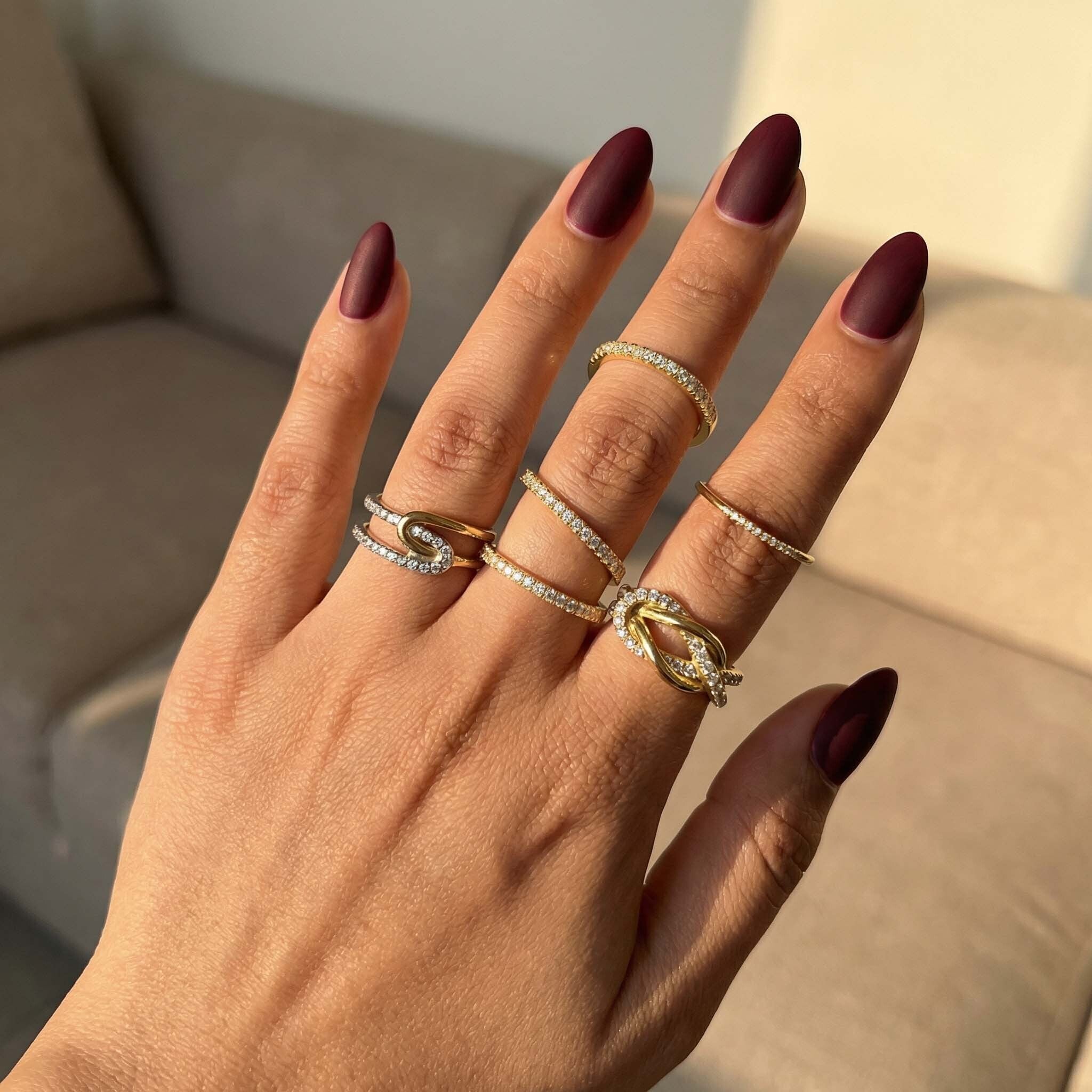 Hand with maroon nail polish wearing multiple gold rings on a neutral background