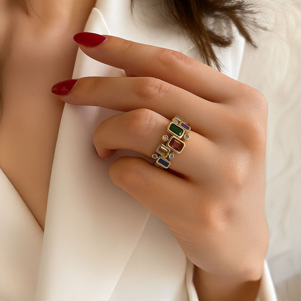 Close-up of a hand wearing a gold ring with colored gemstones on a light background