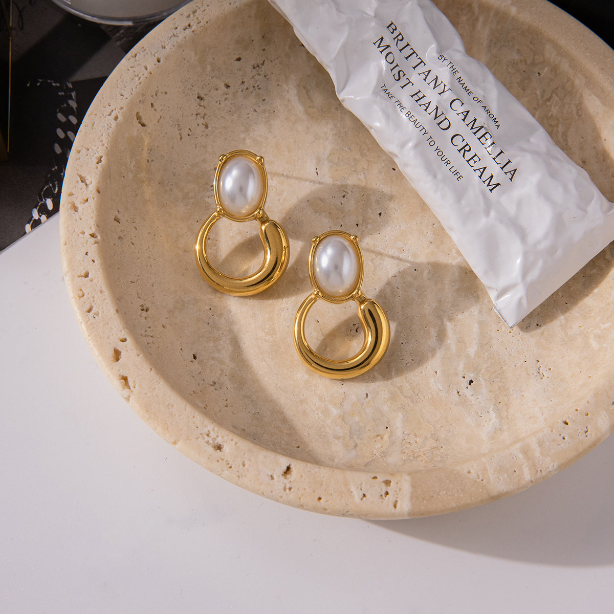 Gold earrings for women with pearl-like stones on a beige stone surface next to a white hand cream tube.