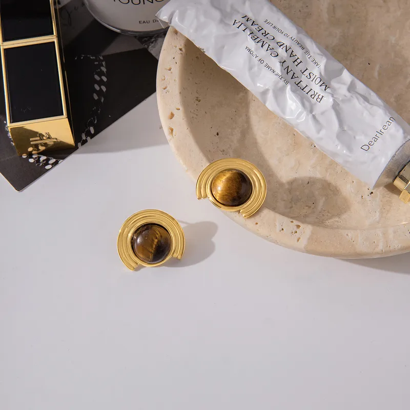 Gold earrings for women on a white surface with a beige plate and product packaging in the background.