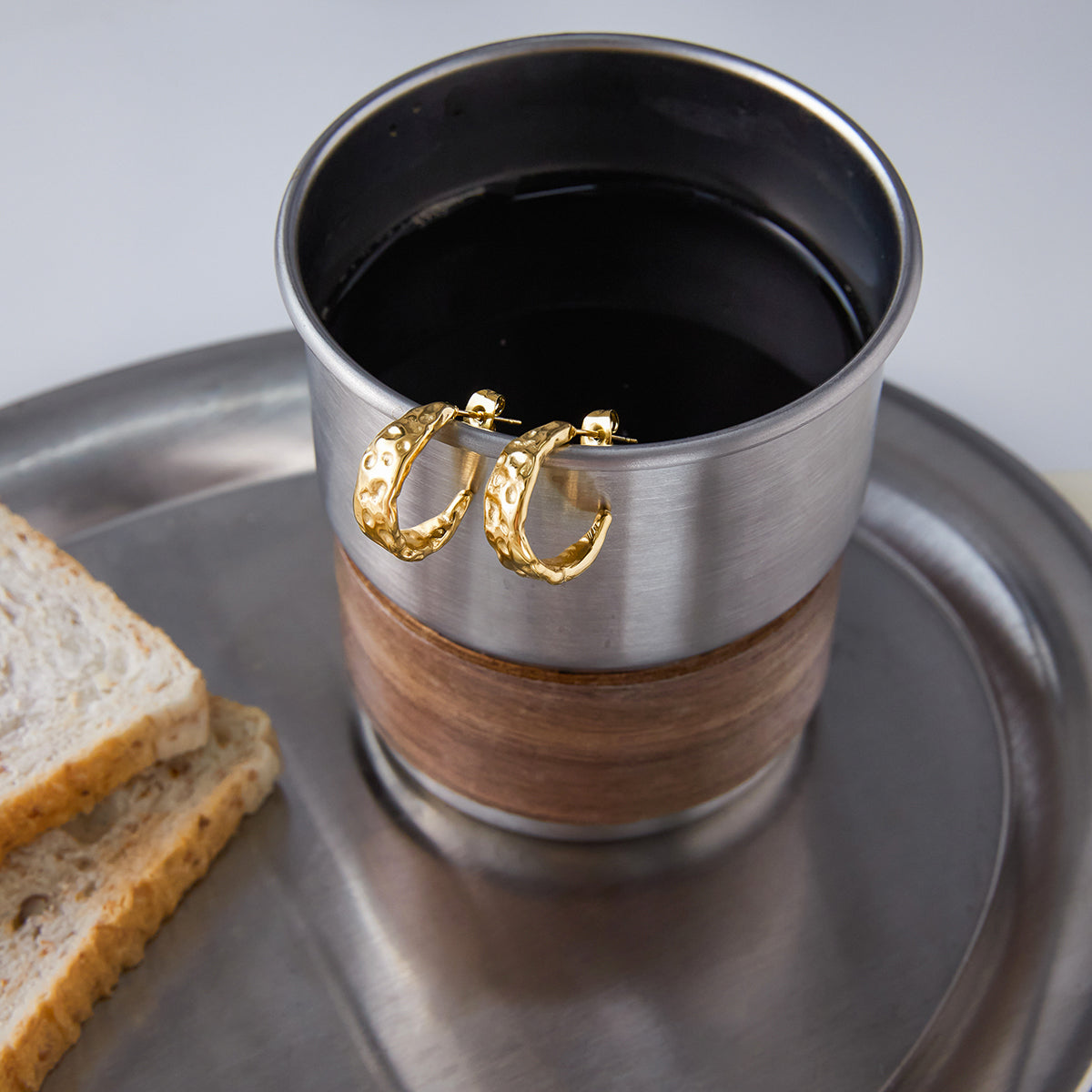 Silver and wooden mug with gold hoop earrings on a metal tray with bread slices.
