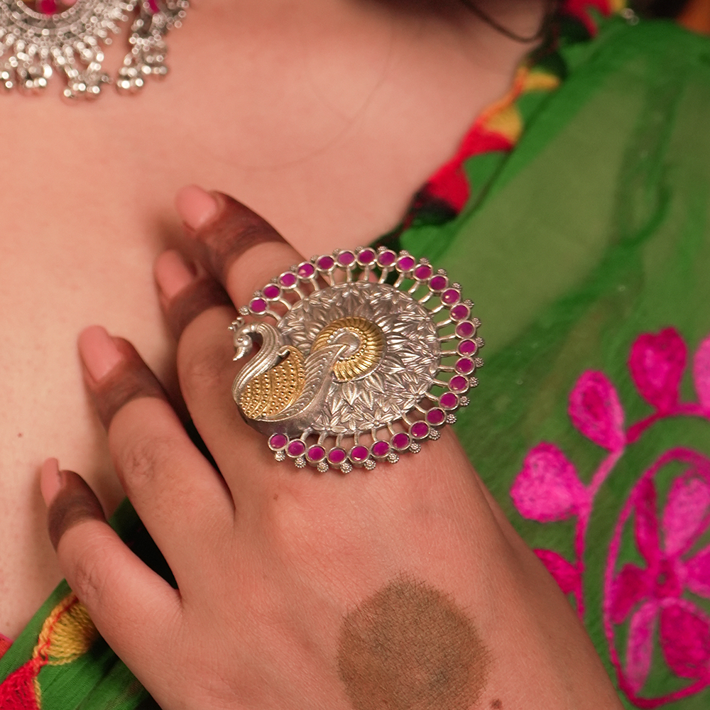 Decorative ring with peacock design worn on a hand against a colorful traditional outfit.