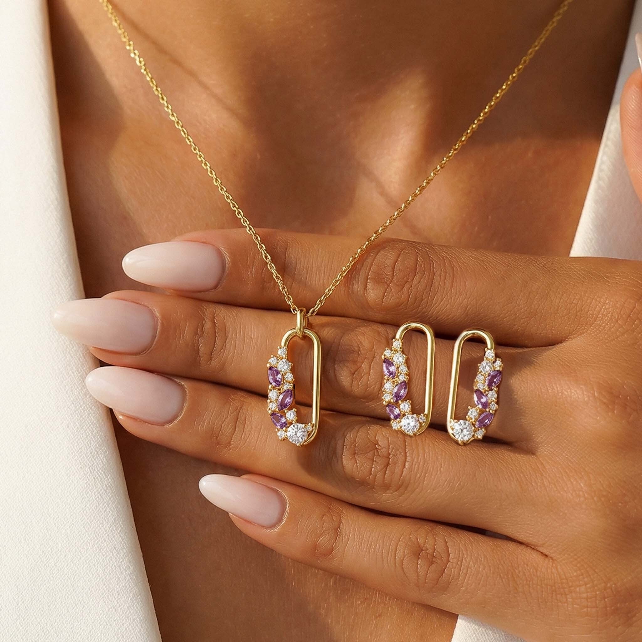 Gold necklace with purple and white gemstones held by a hand with pink nail polish.