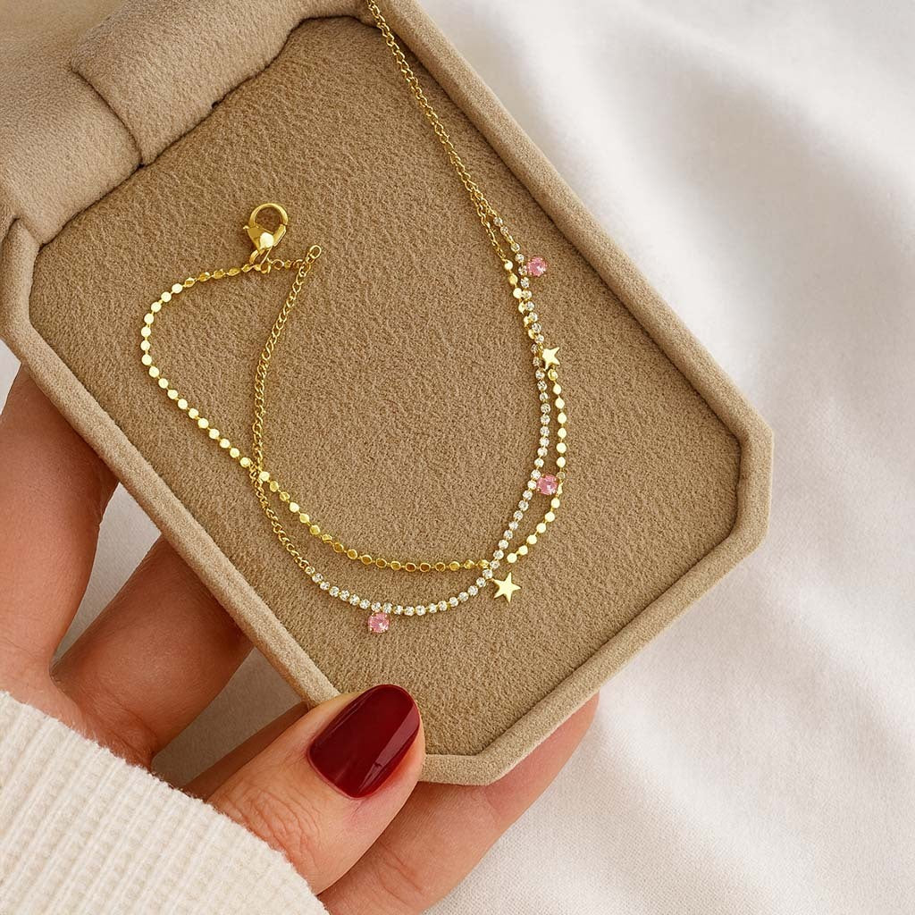 Gold anklet with star and bead details on a beige jewelry box held by a hand with red nail polish.