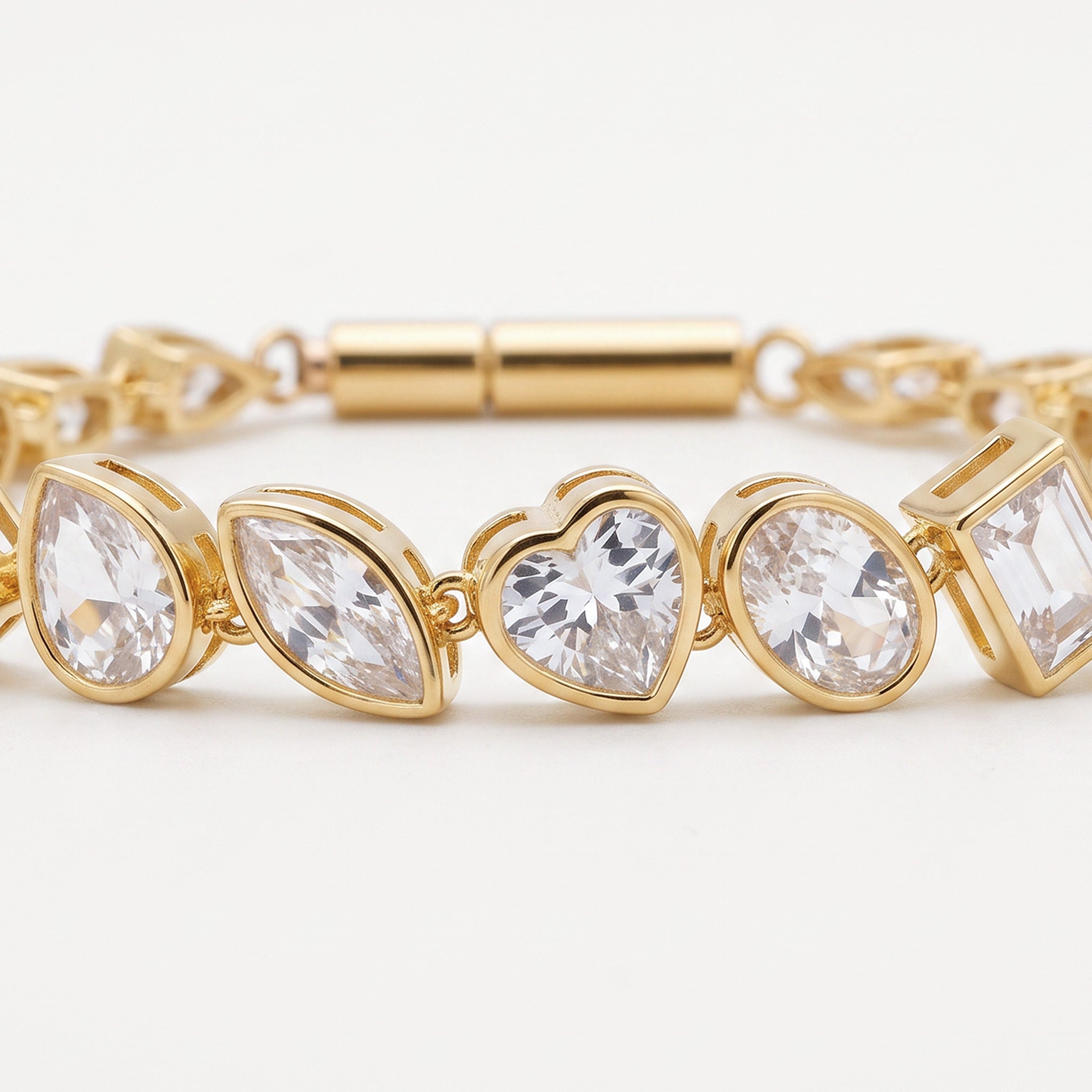 A gold-plated bracelet featuring a row of clear, faceted gemstones in marquise, heart, oval, and square shapes, displayed on a clean white background.