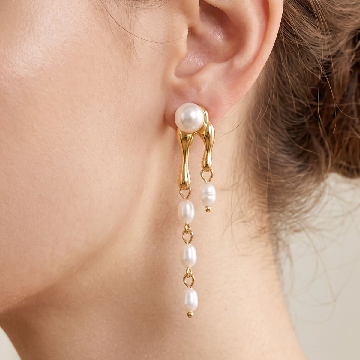 A close-up of a model's ear wearing a gold and pearl drop earring. The earring features a gold huggie-style clasp with a white pearl at the top, and three dangling white pearls connected by small gold links.