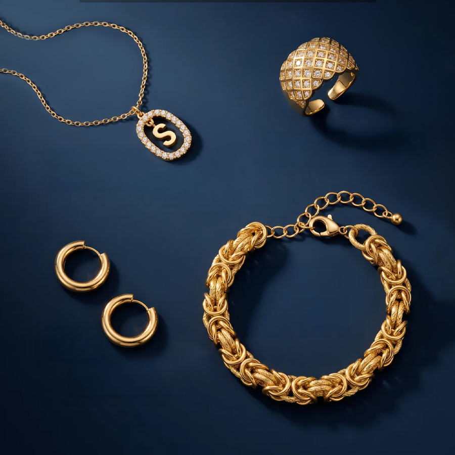 A gold jewelry set arranged on a dark blue background, including a chain necklace with an oval pendant, a textured spherical ring, a braided bracelet, and two hoop earrings.