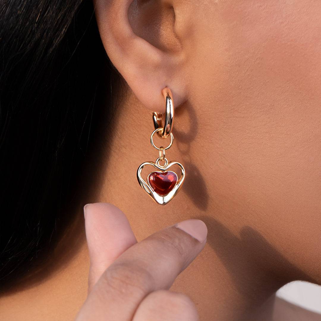 Gold heart-shaped earring with a red gemstone worn by a person.