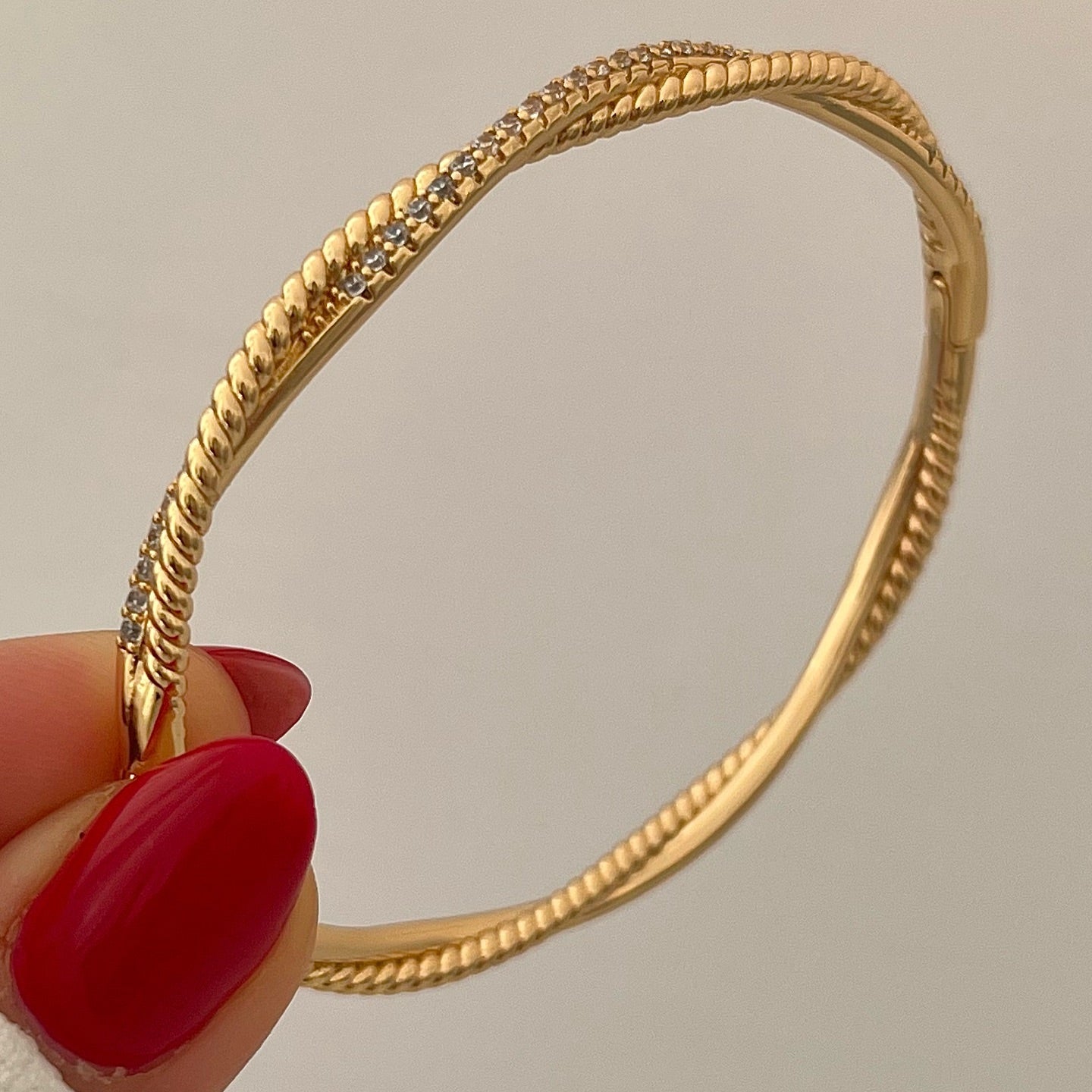 Gold bracelet held by a hand with red nail polish against a neutral background