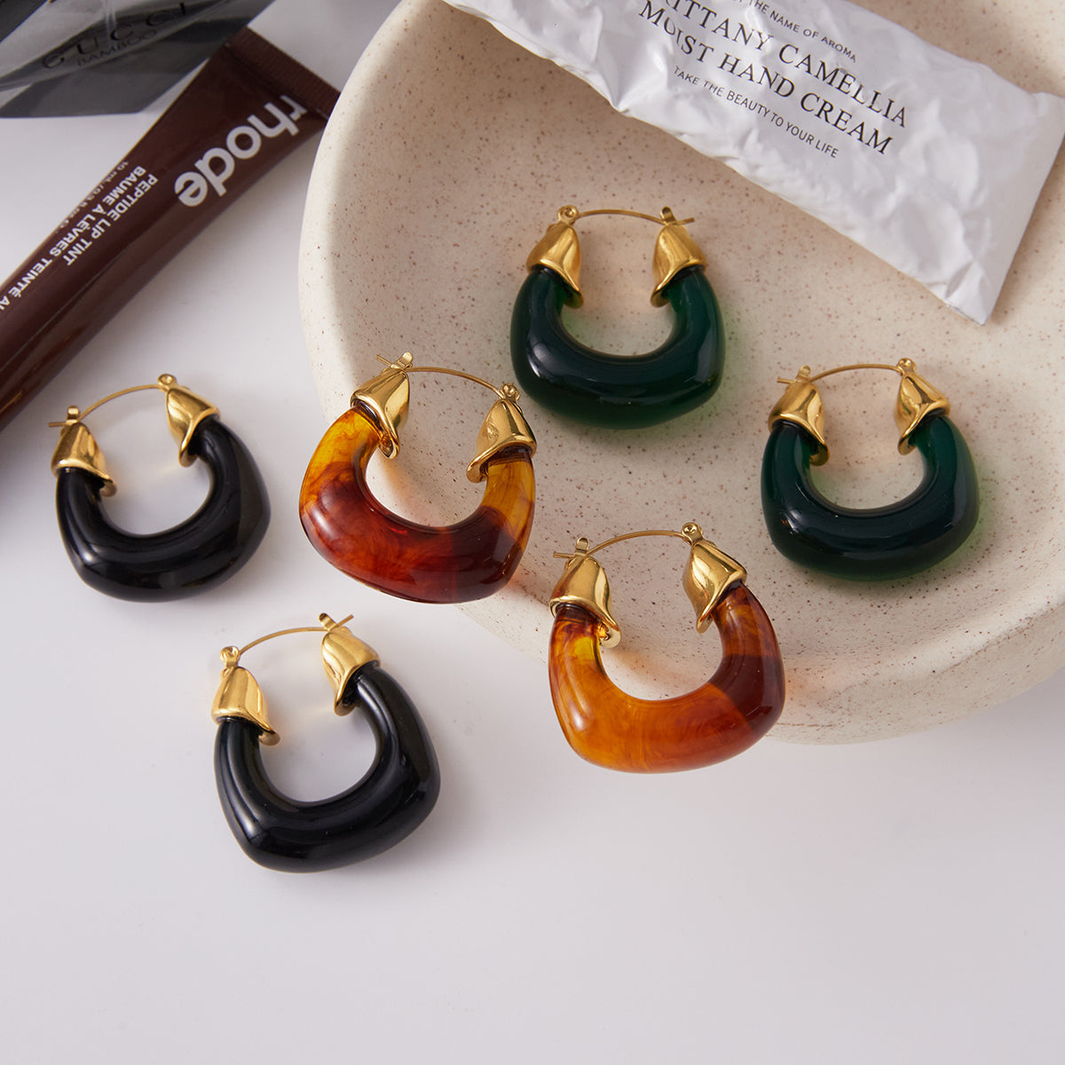 Set of hoop earrings in black, brown, and green on a textured surface with a hand cream container in the background.