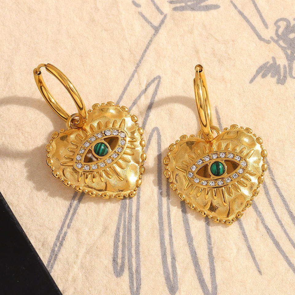 Gold heart-shaped earrings with green gemstones on a textured beige background