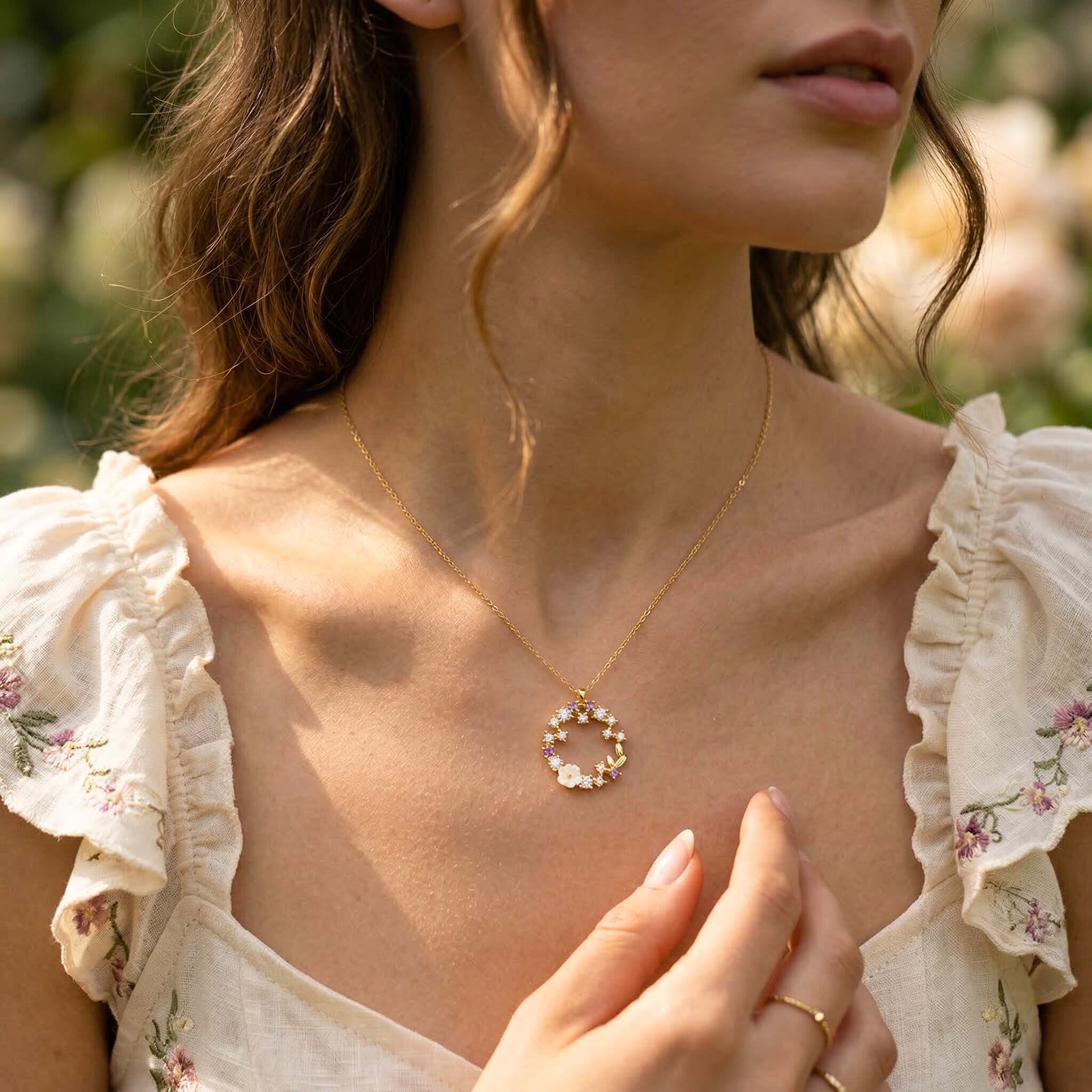 Woman wearing a delicate gold necklace with a floral pendant, set against a blurred natural background.