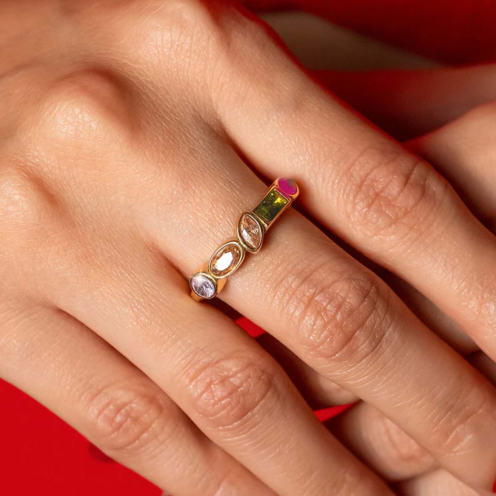 Close-up of a hand wearing a gold ring with colorful gemstones against a red background