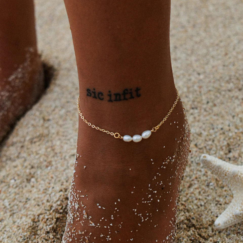 Ankle with gold anklet and 'sic infit' tattoo on sand background