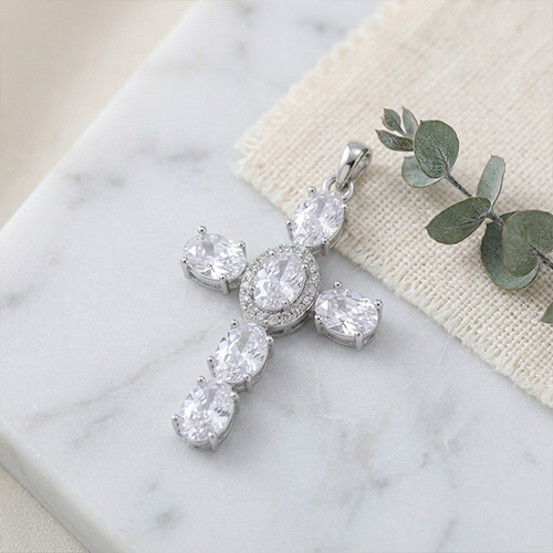 A silver cross Charm with clear oval gemstones, displayed on a white marble surface next to a textured beige fabric and a green eucalyptus branch.