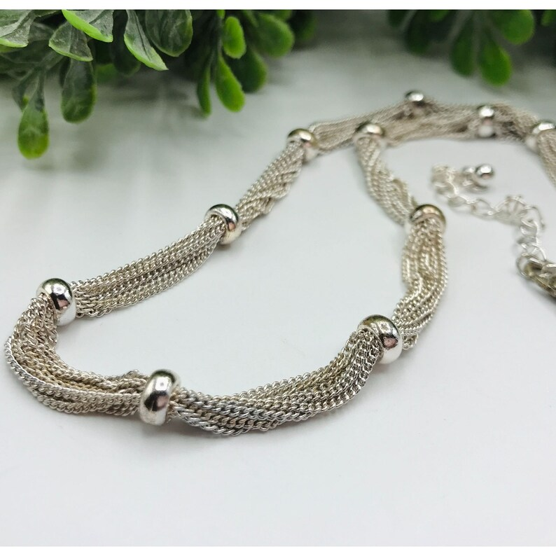 Silver multi-strand necklace on a white surface with green leaves in the background