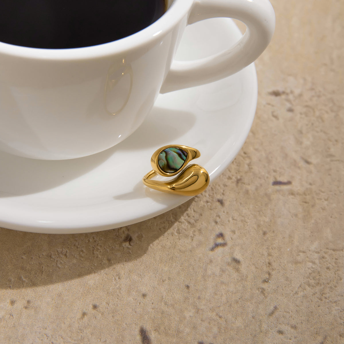 Gold ring with green stone on a white saucer next to a cup of coffee.