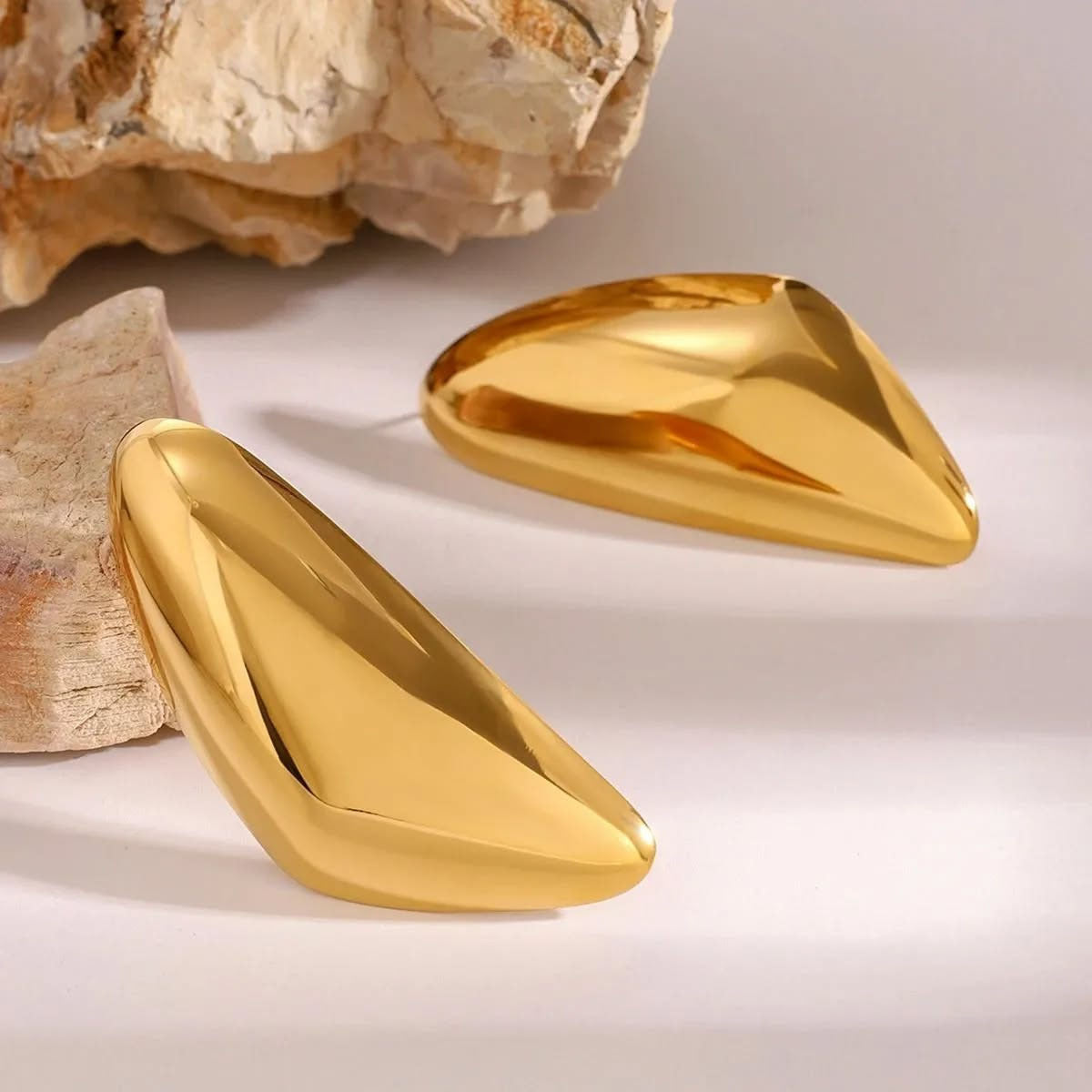 Two gold-colored earrrings for women on a light background with a natural rock texture.