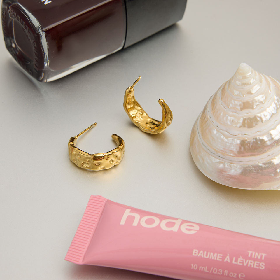 Gold hoop earrings with a pink lip balm labeled 'hode' and a shell on a light surface.