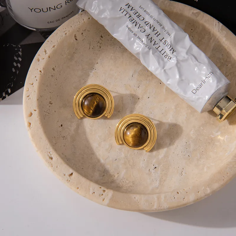 Gold earrings for women on a beige stone surface with a white tube of cream in the background.