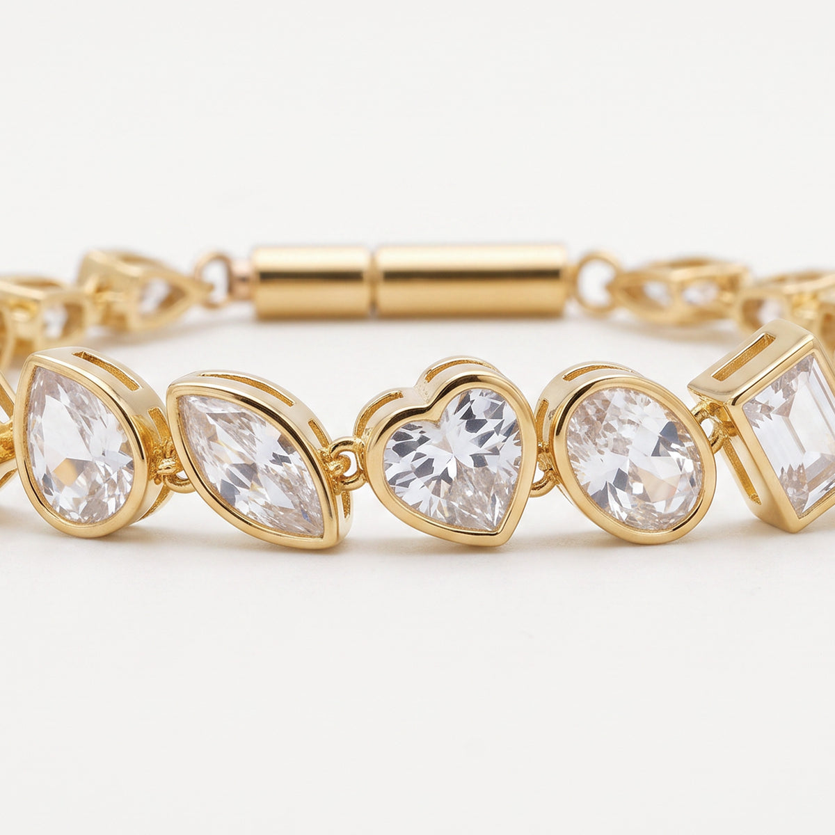 A gold-plated bracelet featuring a row of clear, faceted gemstones in marquise, heart, oval, and square shapes, displayed on a clean white background.