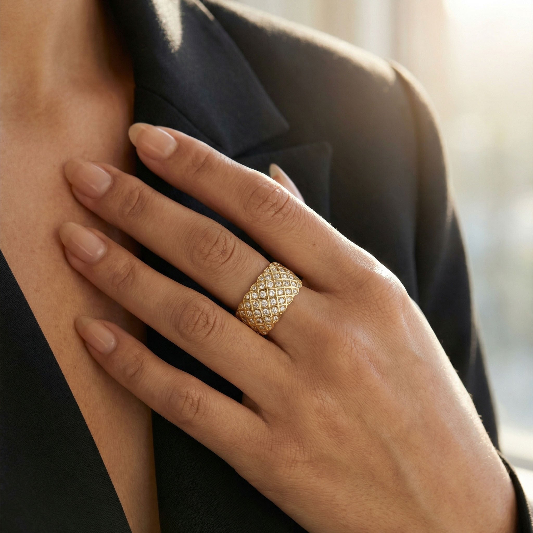 Close-up of a hand wearing a wide gold ring with a diamond lattice pattern, resting against a black blazer.