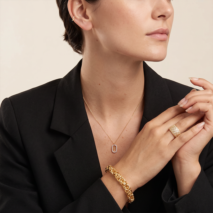 A close-up lifestyle shot of a model wearing a gold chain necklace with a small rectangular pendant, a textured gold ring, and a gold chain bracelet, styled with a black blazer against a warm beige background.