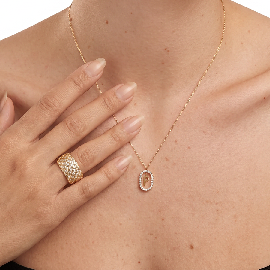 A close-up of a model's neck and chest wearing a delicate gold chain necklace with an oval pendant set with small clear stones, paired with a textured gold ring on the finger.