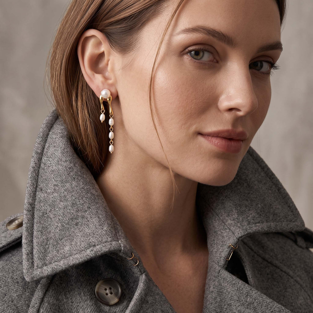 A close-up of a woman wearing a gold and pearl drop earring, styled with a gray wool coat against a neutral textured background.