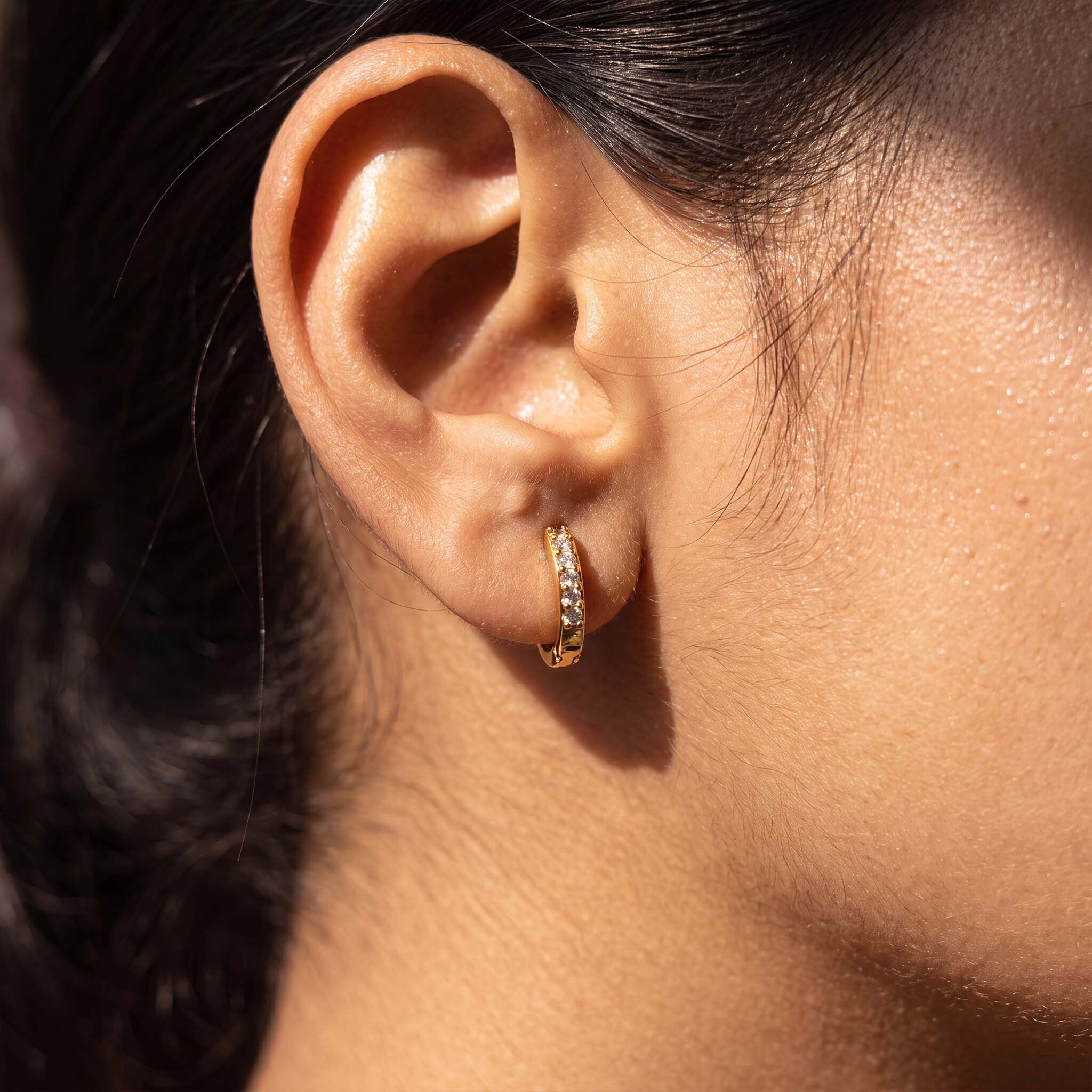 A close-up of a person's ear wearing a small gold huggie hoop earring  with clear stones, shown in bright natural sunlight.