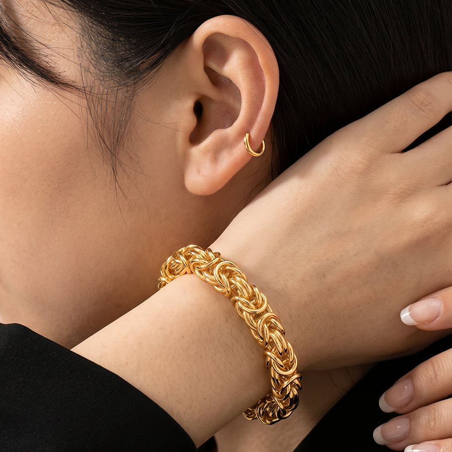textured gold chain link bracelet on her wrist and a small gold hoop earring in her ear. She is wearing a black top, and her hand rests near her head.