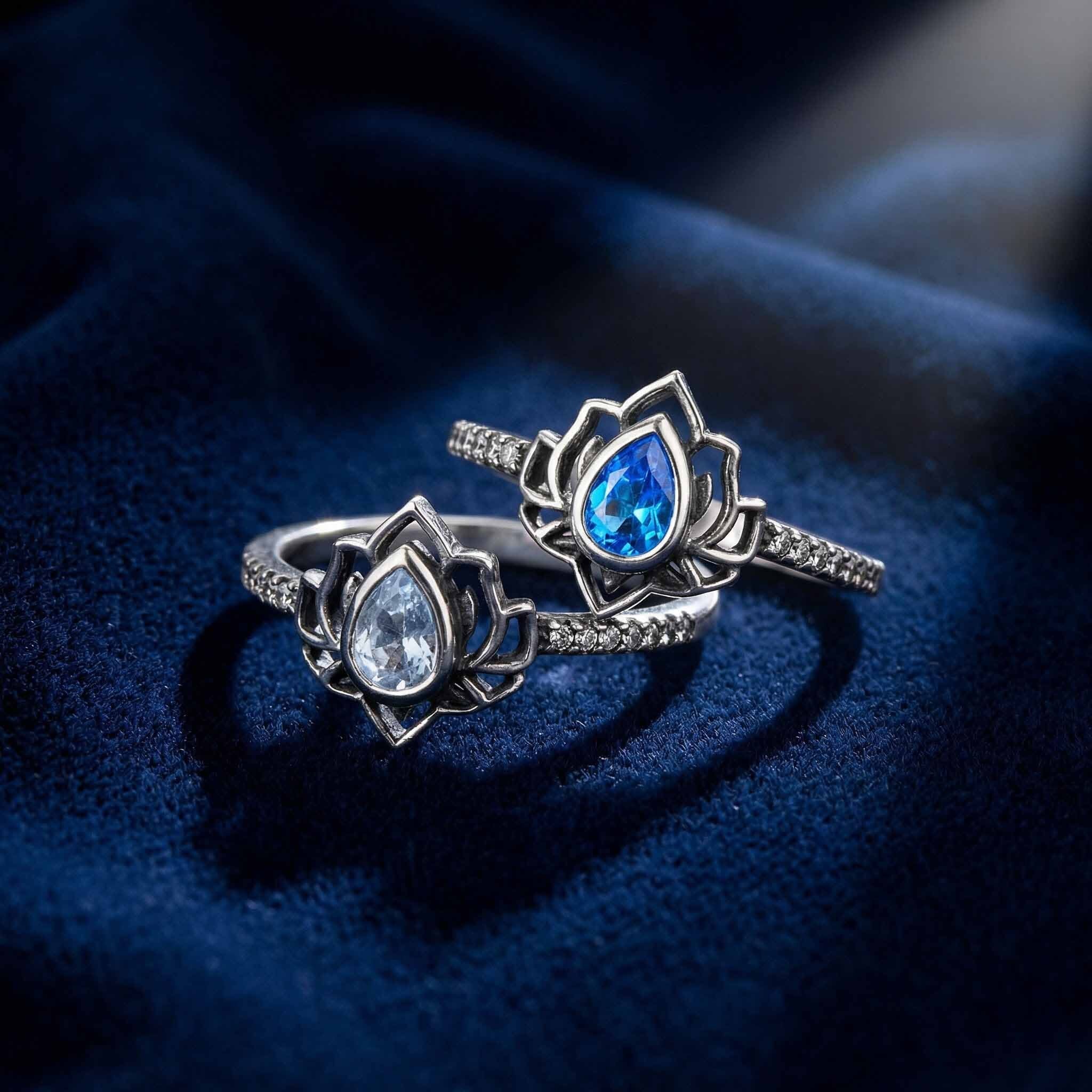 Two silver lotus flower rings with gemstones on a dark blue fabric background